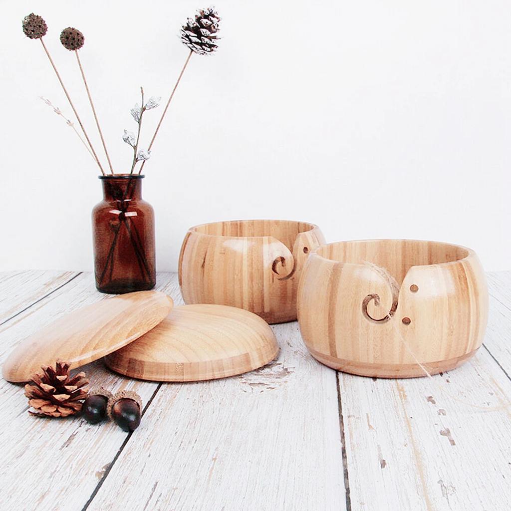 Two Wooden Yarn Bowl With Lid, each round with carved swirl details and a high gloss finish, rest on a white wooden surface next to a brown vase filled with dried plants and several pinecones.