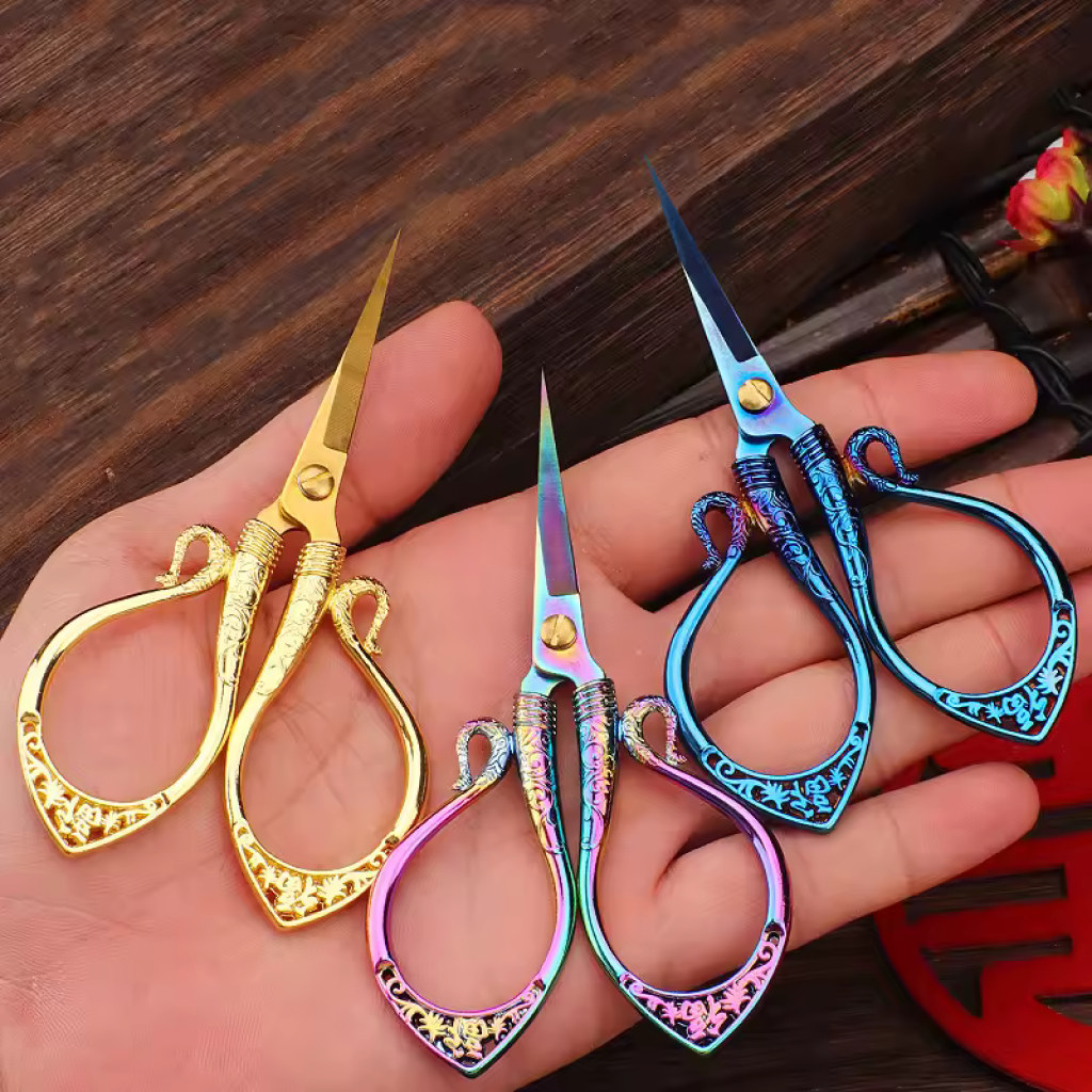 A hand holds three Decorative Vintage Embroidery Scissors in gold, rainbow, and blue on a wooden surface—ideal for precise crafting and bringing charm to your sewing essentials.