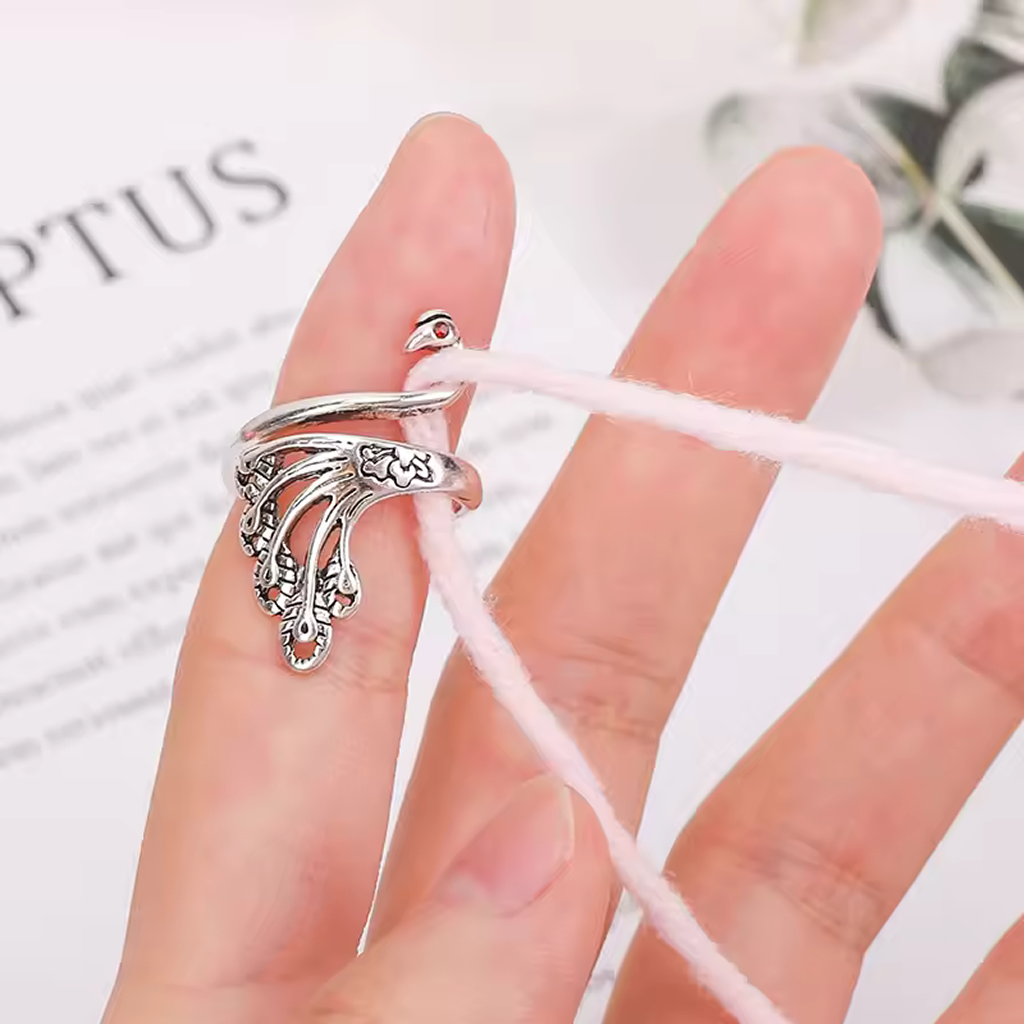 A Yarn Tension Control Ring, featuring an ornate swan and peacock design in silver, is shown worn on a person's finger with a pink yarn looped around the ring and hand.