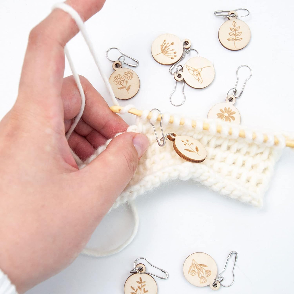 A hand knits with cream-colored yarn, using durable Wooden Flower Stitch Markers with Storage Box 8Pcs engraved with botanical designs. Several markers and the storage box are scattered on the white surface.