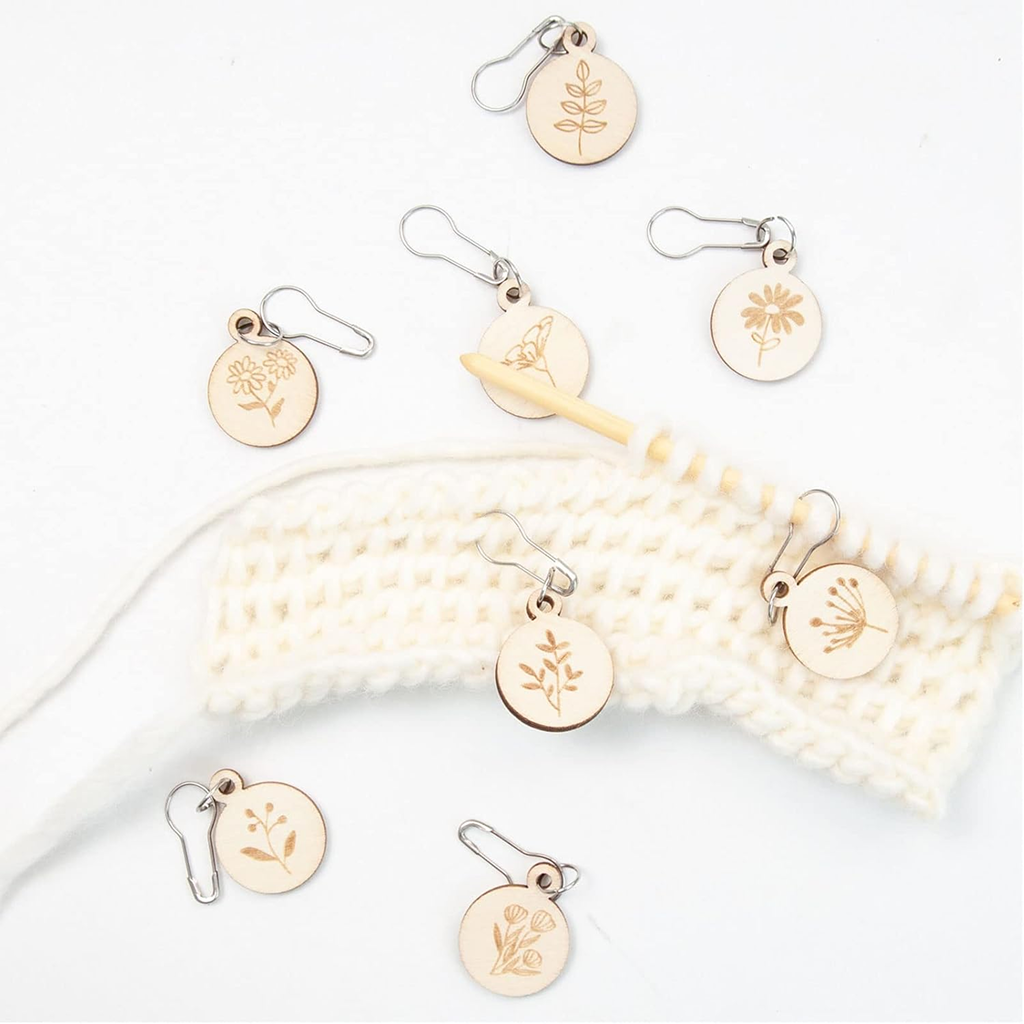 Eight Wooden Flower Stitch Markers with botanical engravings are displayed on a white knitted swatch beside a yellow crochet hook—perfect for adding charm to your crochet projects. Set includes 8 pieces and a handy storage box.