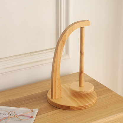 A wooden headphone stand with a curved support and round base sits on a wooden surface beside eyeglasses, a magazine, and the Levitating Premium Yarn Holder for seamless knitting.