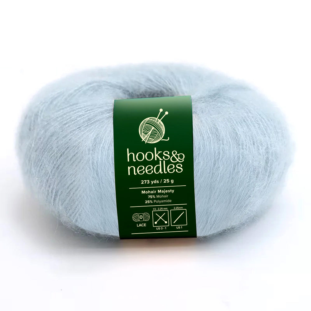 A skein of Mohair Majesty Yarn 25g in light blue with a green label that reads "hooks & needles, 75% mohair, 25% polyamide, 273 yds / 25 g." This yarn's luxurious feel is perfect for any delicate project.