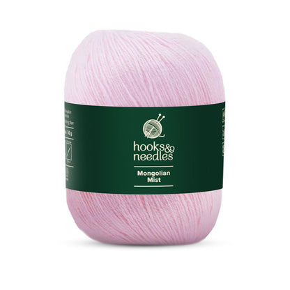 A skein of light pink "Mongolian Mist Yarn 50g" presented on a dark green band with the label "Hooks & Needles," made from super fine-weight yarn for a smooth, luxurious feel.