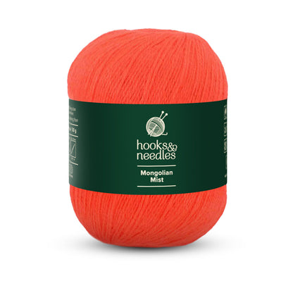 Bright orange yarn, identified as "Mongolian Mist Yarn 50g" under the "hooks & needles" label, is wrapped in green and crafted from super fine-weight Mongolian Cashmere. This anti-pilling fiber keeps your creations vibrant and smooth over time.