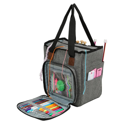 The Yarn Organizer Tote in gray, complete with black straps, is packed with knitting tools, colorful pens, and yarn. It also has a front transparent pocket that is ideal for storing crochet hooks.