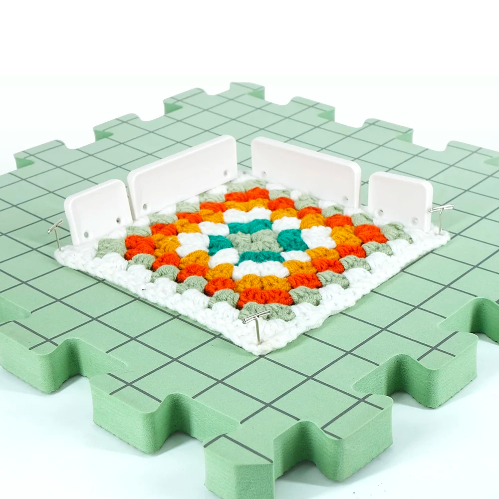 A colorful crocheted square is pinned and drying on a Knitting Crocheting Blocking Mat, which features a convenient grid pattern, with the backdrop of a green foam material.