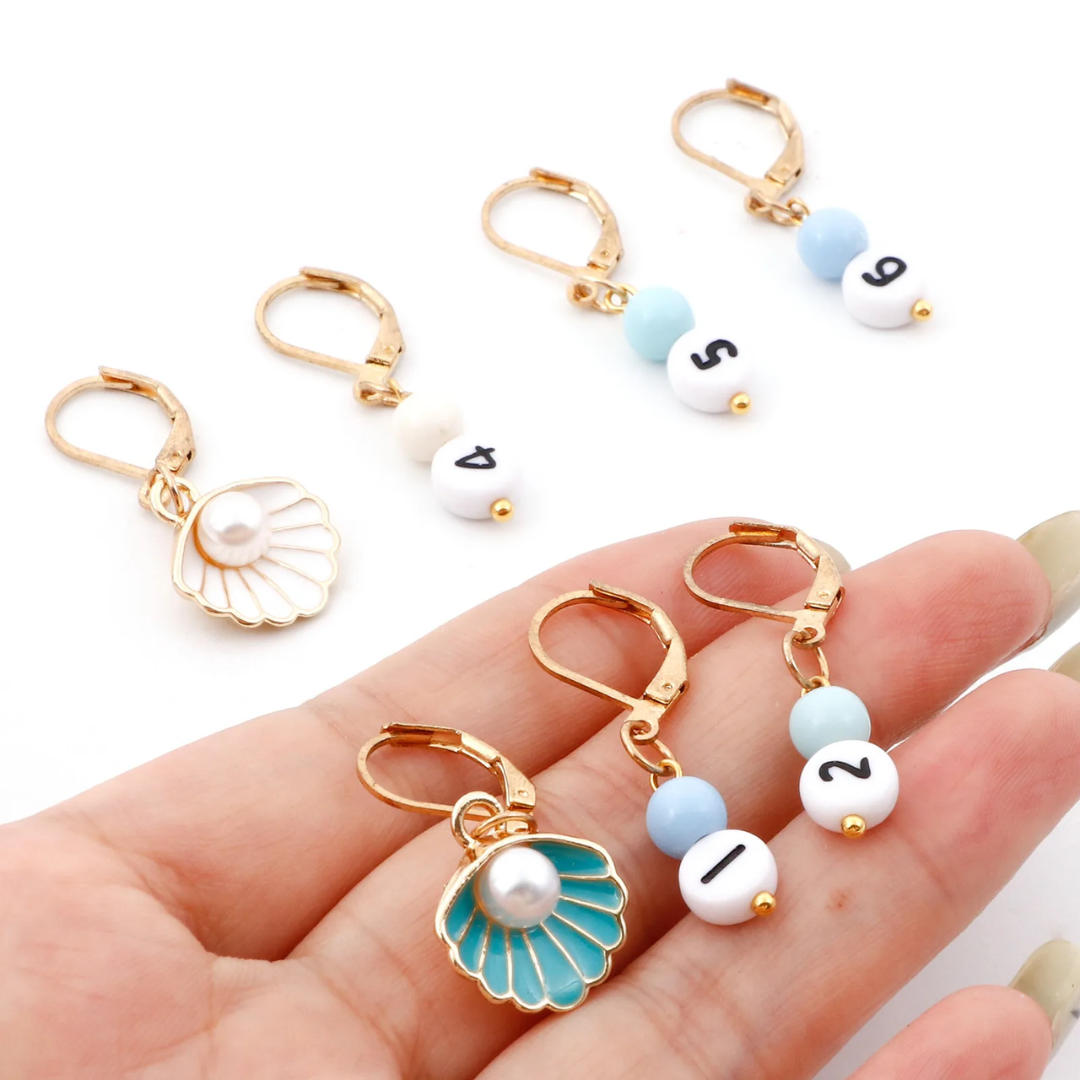 Hand holding the 35-Piece Knitting Kit: Perfect for Beginners, featuring golden stitch markers with numbers and charming shell designs, as well as additional numbered stitch markers, ideal for those looking to enhance their knitting tools collection, displayed on a white background.