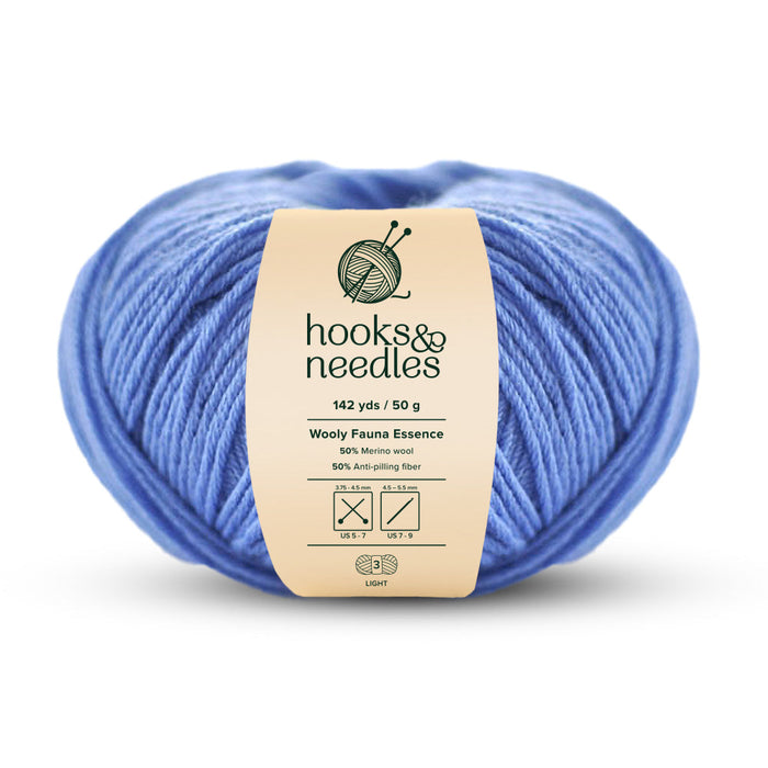 A skein of Wooly Fauna Essence Yarn 50g in light blue, labeled "hooks & needles," provides 142 yards/50 grams of merino wool blended with anti-pilling fiber, ideal for US 5-7 needles.