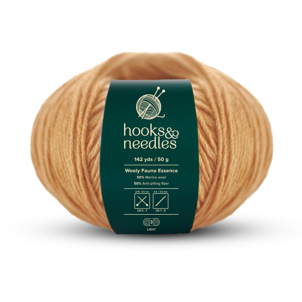 A light brown ball of Wooly Fauna Essence Yarn 50g, labeled "hooks & needles," provides 142 yards of luxurious merino wool blended with anti-pilling fiber for a smooth crafting experience.