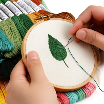 A person uses green threads from the Embroidery Floss Set to stitch a leaf on white fabric in a wooden hoop, with other colorful flosses nearby.