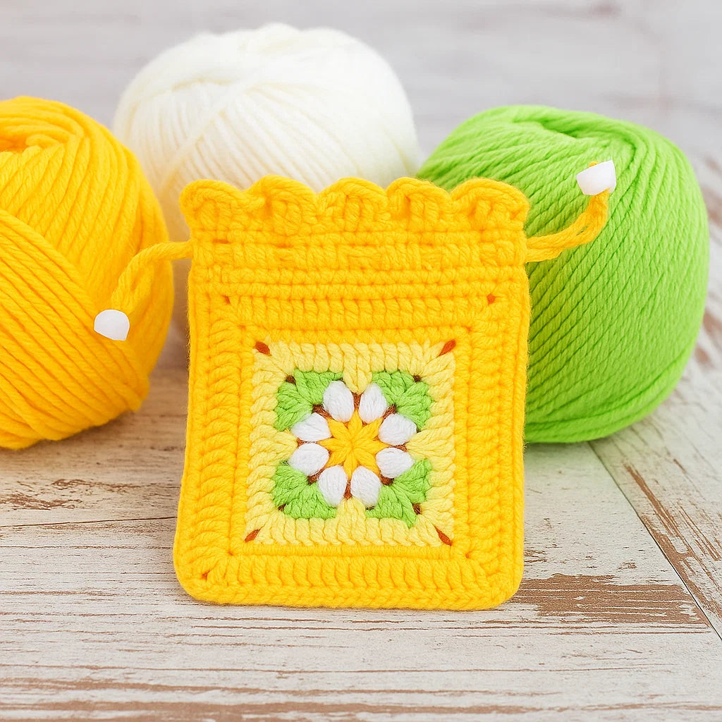 A small crocheted drawstring pouch, made with the Crochet Flower Purse Kit, featuring a yellow, green, and white floral design is displayed in front of three matching yarn balls.