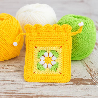 A small crocheted drawstring pouch, made with the Crochet Flower Purse Kit, featuring a yellow, green, and white floral design is displayed in front of three matching yarn balls.