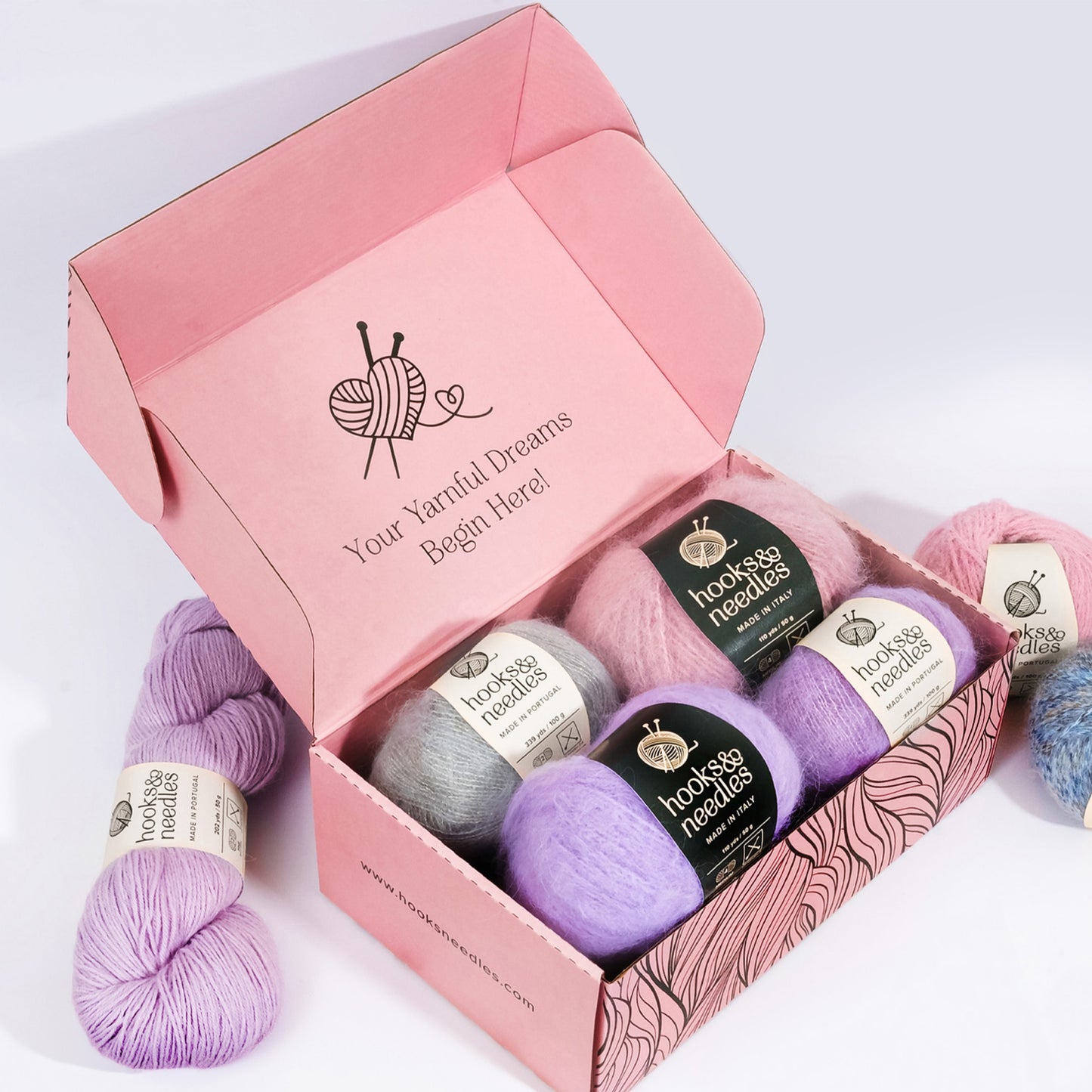 A pink box containing five balls of colored yarn (gray, pink, lavender) from "Hooks & Needles Only Yarn Subscription Box" brand, with two additional balls of yarn outside the box. The box features a knitting-themed design.