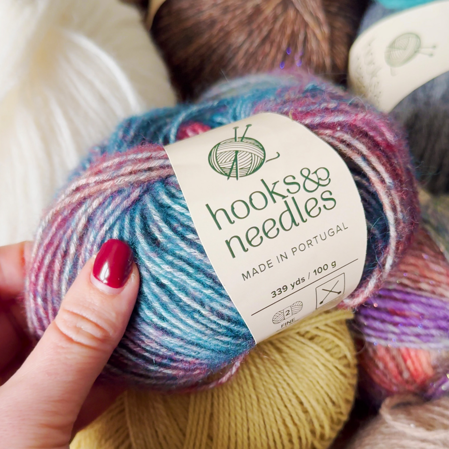 A hand holding a skein of multicolored yarn with a label that reads "[12-Month-Prepaid] Hooks & Needles Subscription Box #30, made in Portugal.