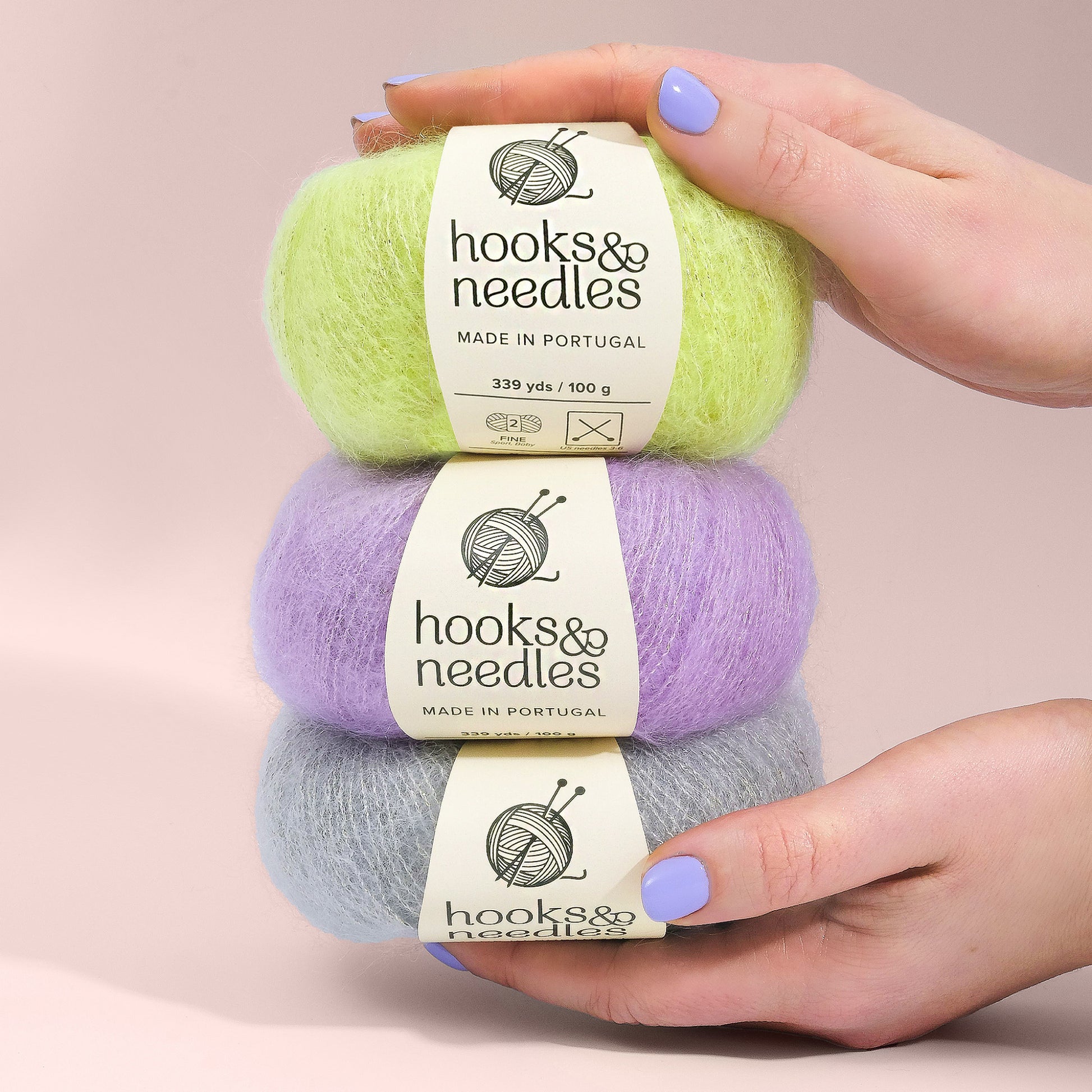 Two hands holding three balls of yarn in green, purple, and gray colors, labeled "Hooks & Needles Only Yarn Subscription Box," made in Portugal.