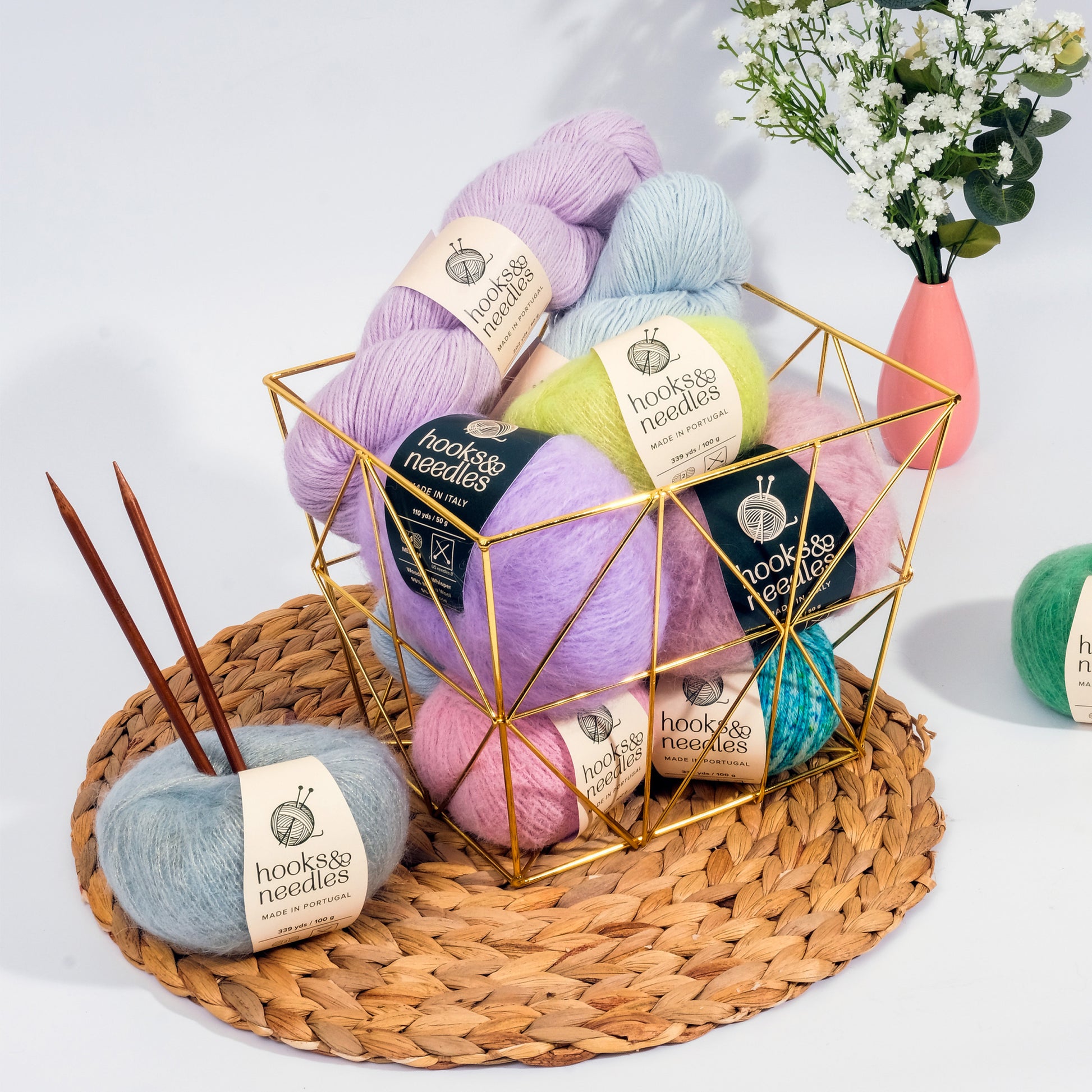 A gold wire basket filled with colorful skeins of yarn on a woven mat. Two wooden knitting needles are inserted into a ball of gray yarn labeled "Hooks & Needles Only Yarn Subscription Box." A vase with flowers is in the background.