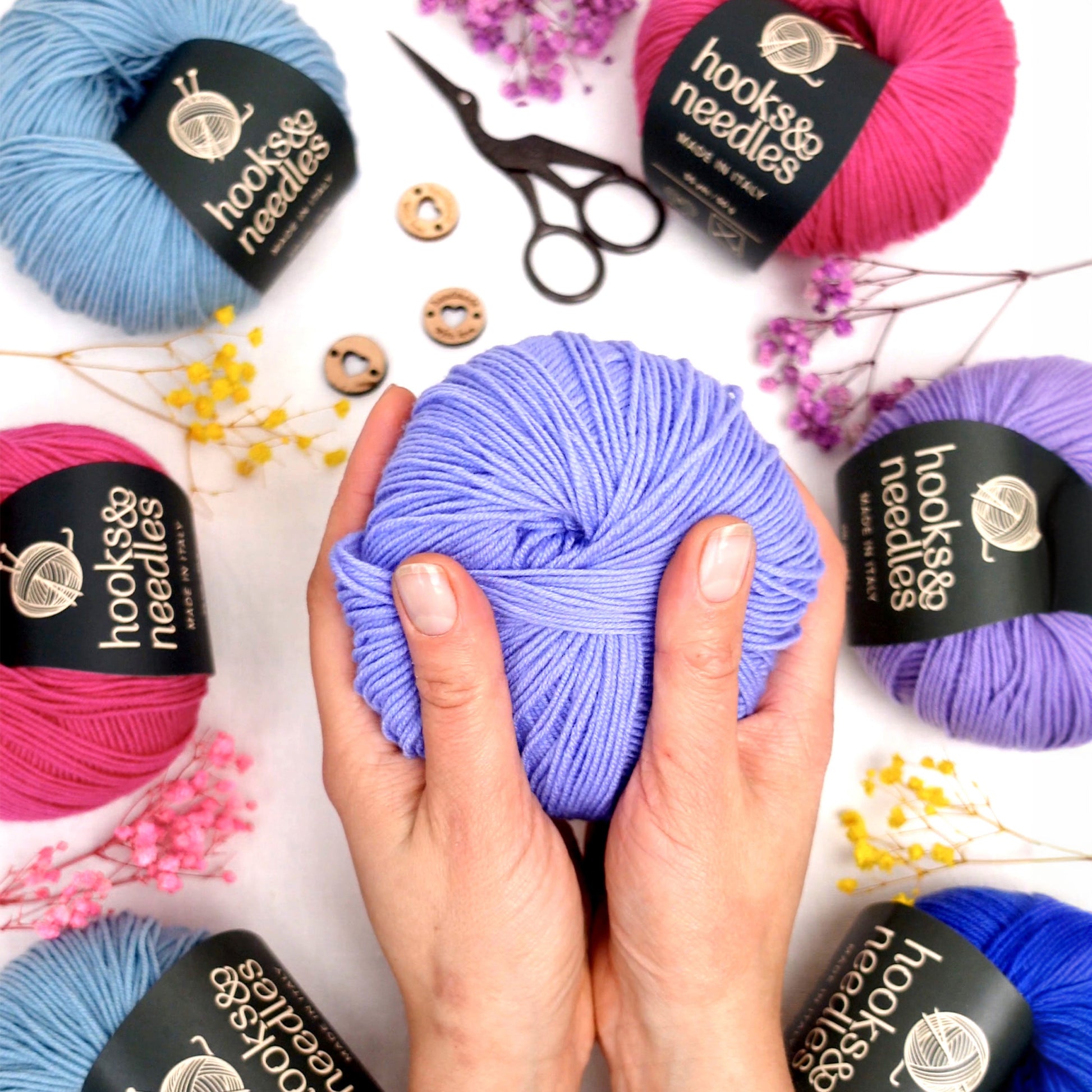 Hands holding a ball of purple yarn surrounded by other balls of yarn, scissors, buttons, and dried flowers from the Hooks & Needles Only Yarn Subscription Box.