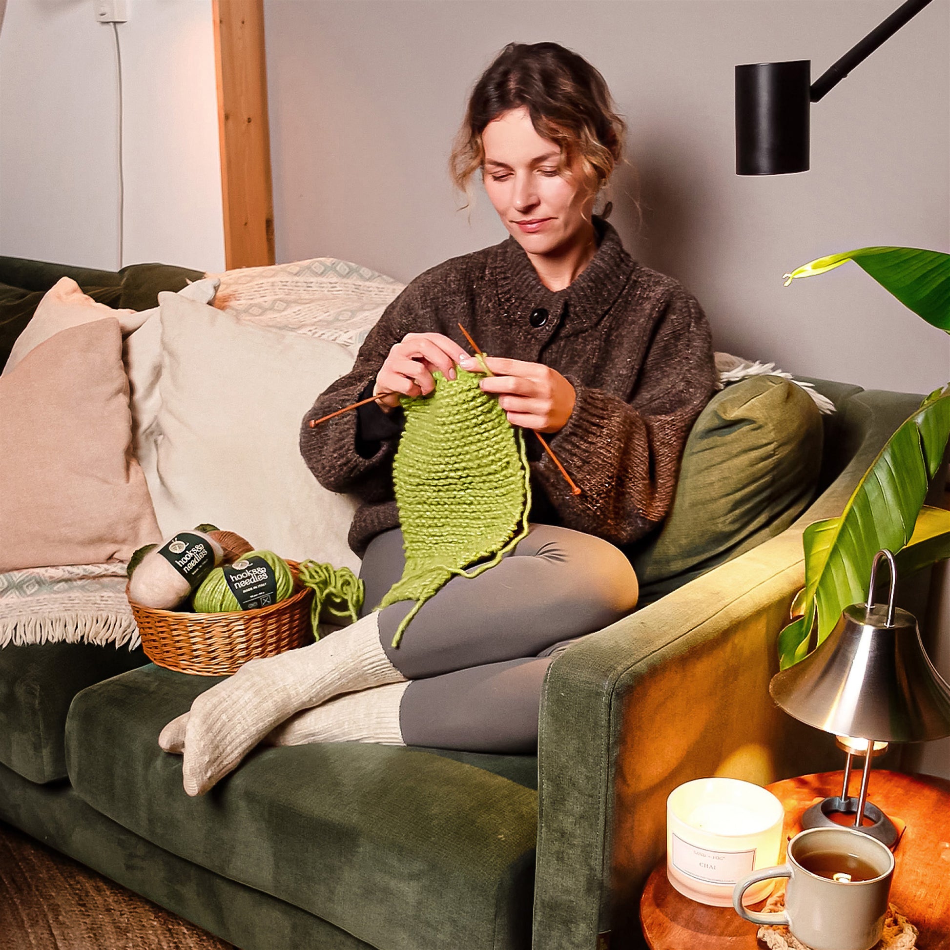 A person sitting on a sofa and knitting a green item. The sofa has pillows, and there is a Hooks & Needles Only Yarn Subscription Box beside them. A coffee cup, candle, and lamp are on a nearby table.