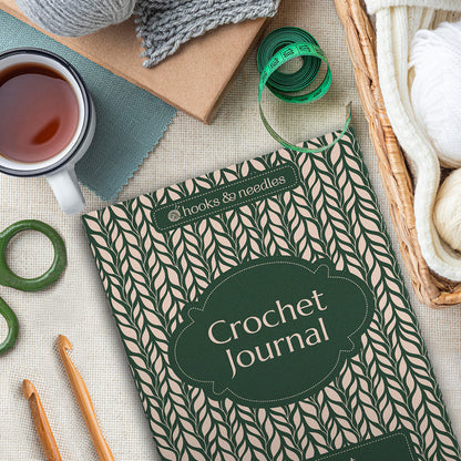 A Crochet Journal is placed on a table with a cup of tea, yarn, green measuring tape, and crochet hooks nearby.