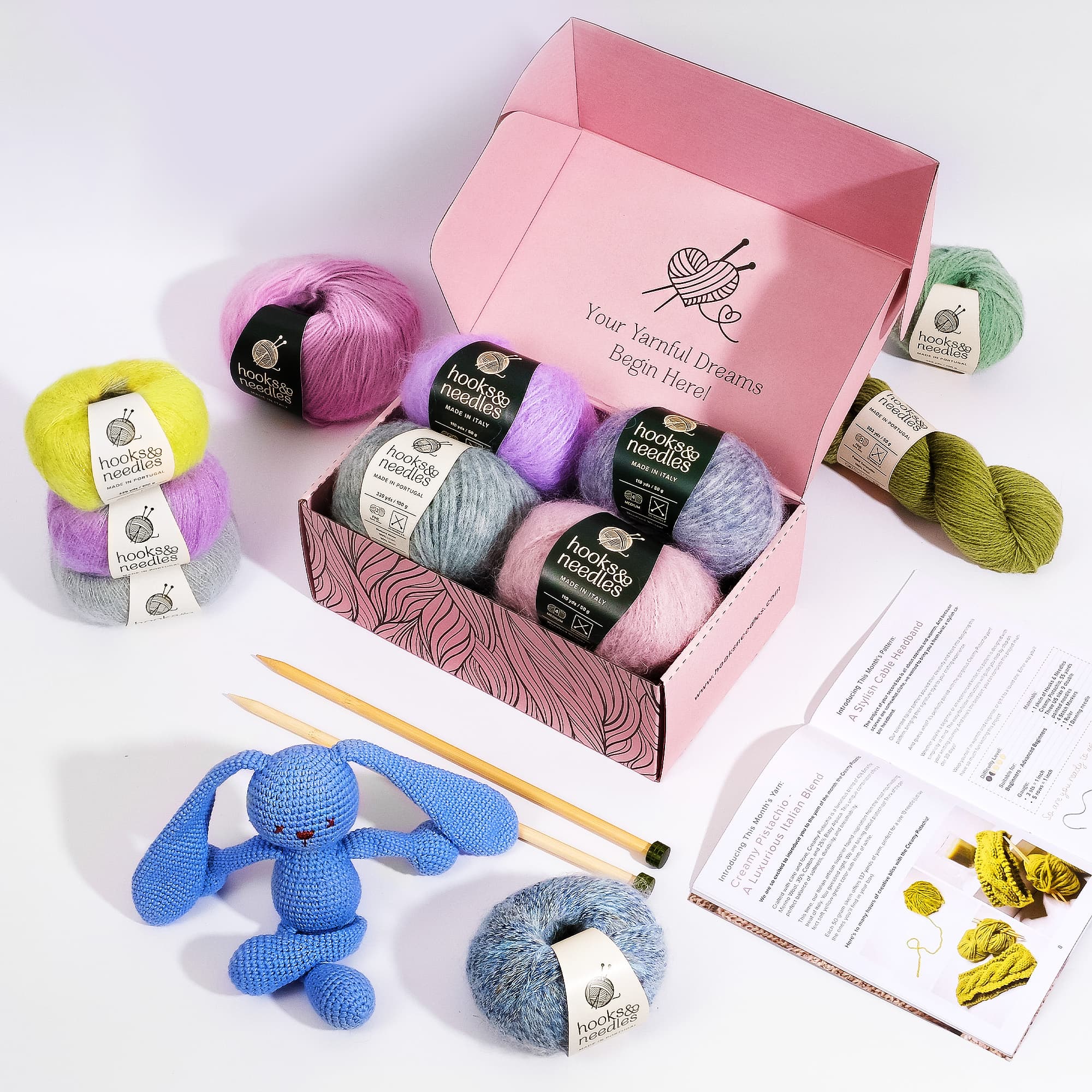 A Hooks & Needles Subscription Box featuring an assortment of colorful yarns, bamboo knitting needles, a pattern booklet, and a finished blue knitted bunny toy.