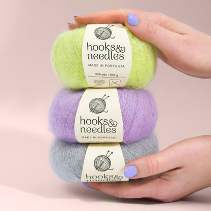 Three Hooks & Needles Subscription Boxes in green, purple, and grey, balanced on each other and held by a hand with blue nail polish.