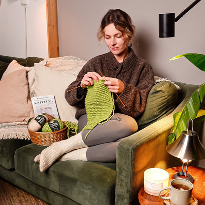 Woman knitting on a couch with Hooks & Needles Subscription Box beside her.