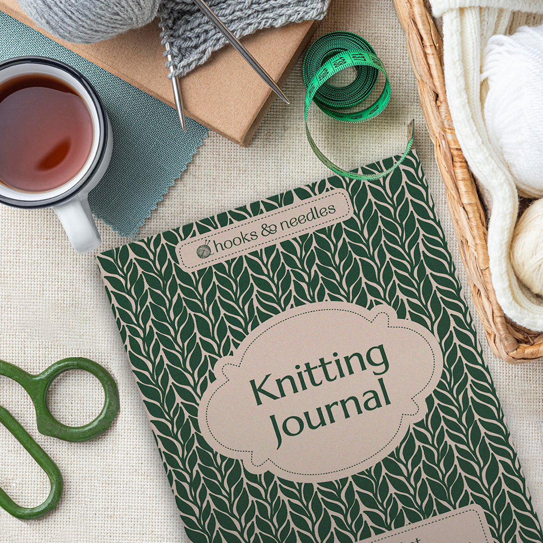 A Knitting Journal, yarn, knitting needles, tape measure, scissors, and a mug of tea are arranged on a light-colored surface.