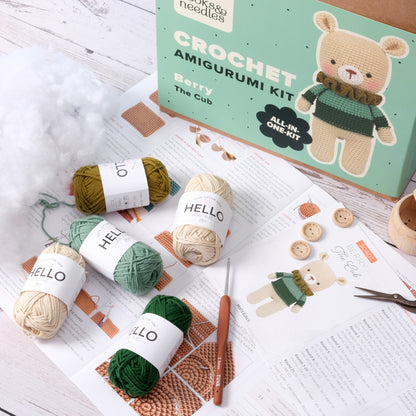 A crochet amigurumi kit for making "Berry the Cub" with yarn, stuffing, a crochet hook, buttons, and instruction sheets shown on a table has been replaced with the Amigurumi Subscription Box.