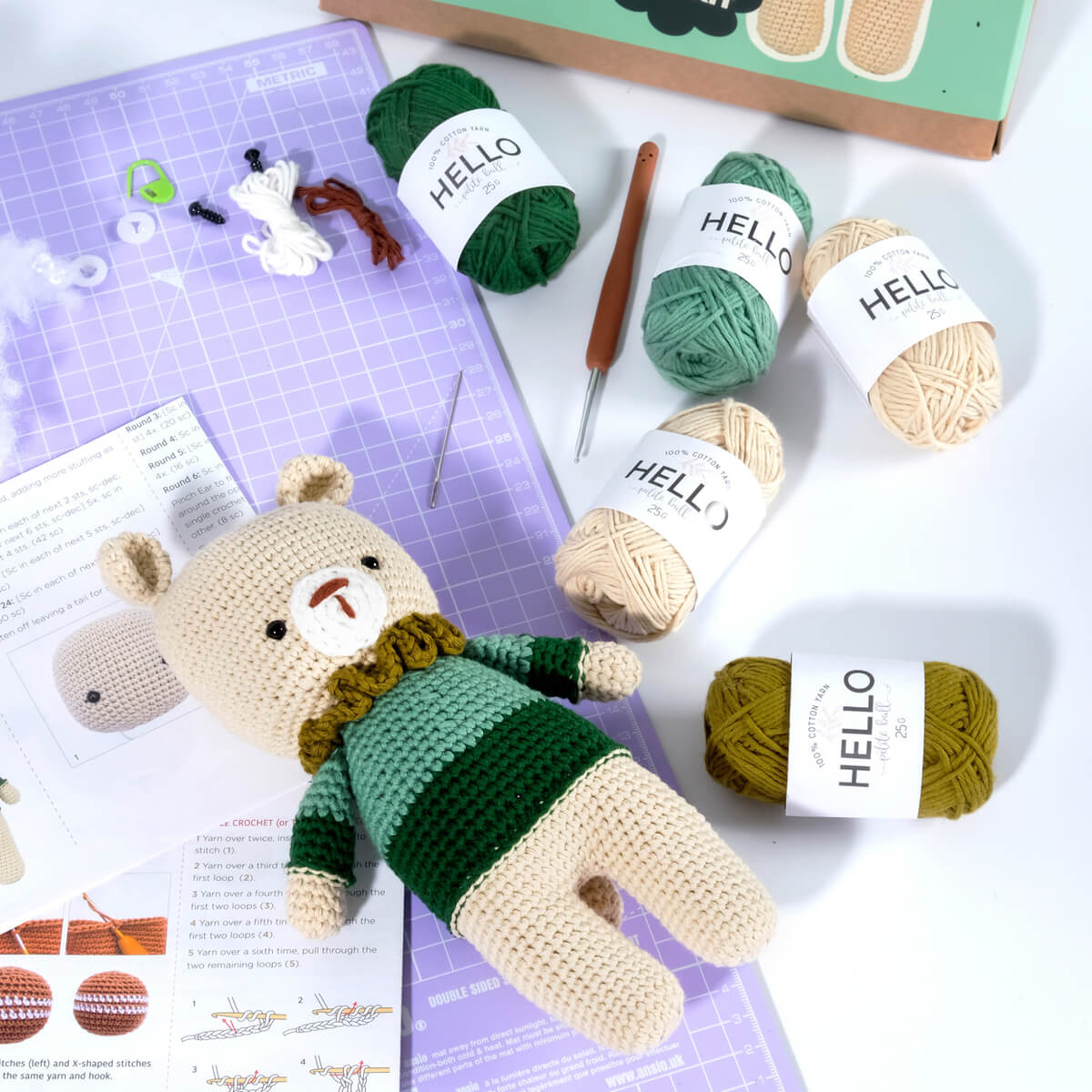 The Amigurumi Subscription Box is displayed with a completed crochet bear resting on a purple crafting mat. The background features yarn in various shades of green and beige, alongside a crochet hook and instruction sheets.