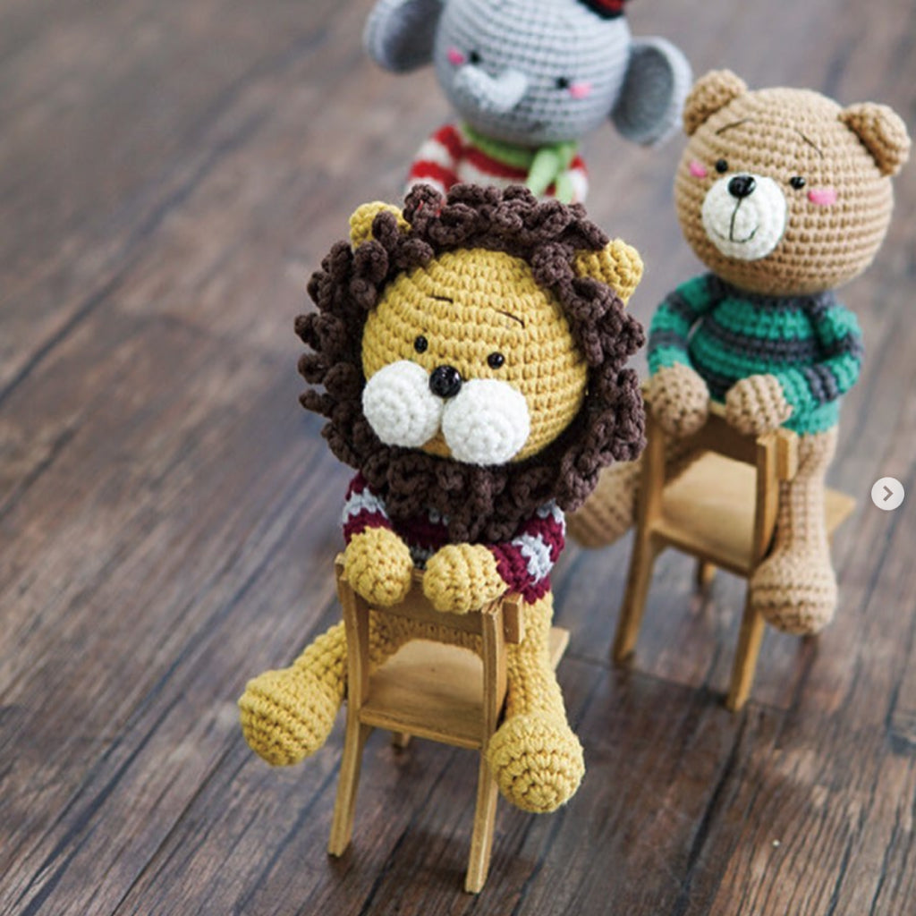 Three Amigurumi Subscription Box items, including a lion, bear, and elephant, are seated on small wooden chairs arranged in a line on a wooden floor.