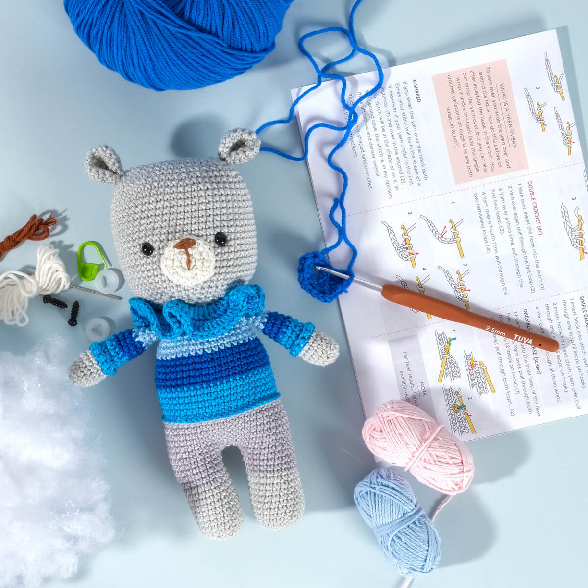 An "Amigurumi Subscription Box" featuring a crocheted bear made with blue and gray yarn, along with various crochet supplies such as a hook, stuffing, and an instruction booklet, arranged on a light blue surface.