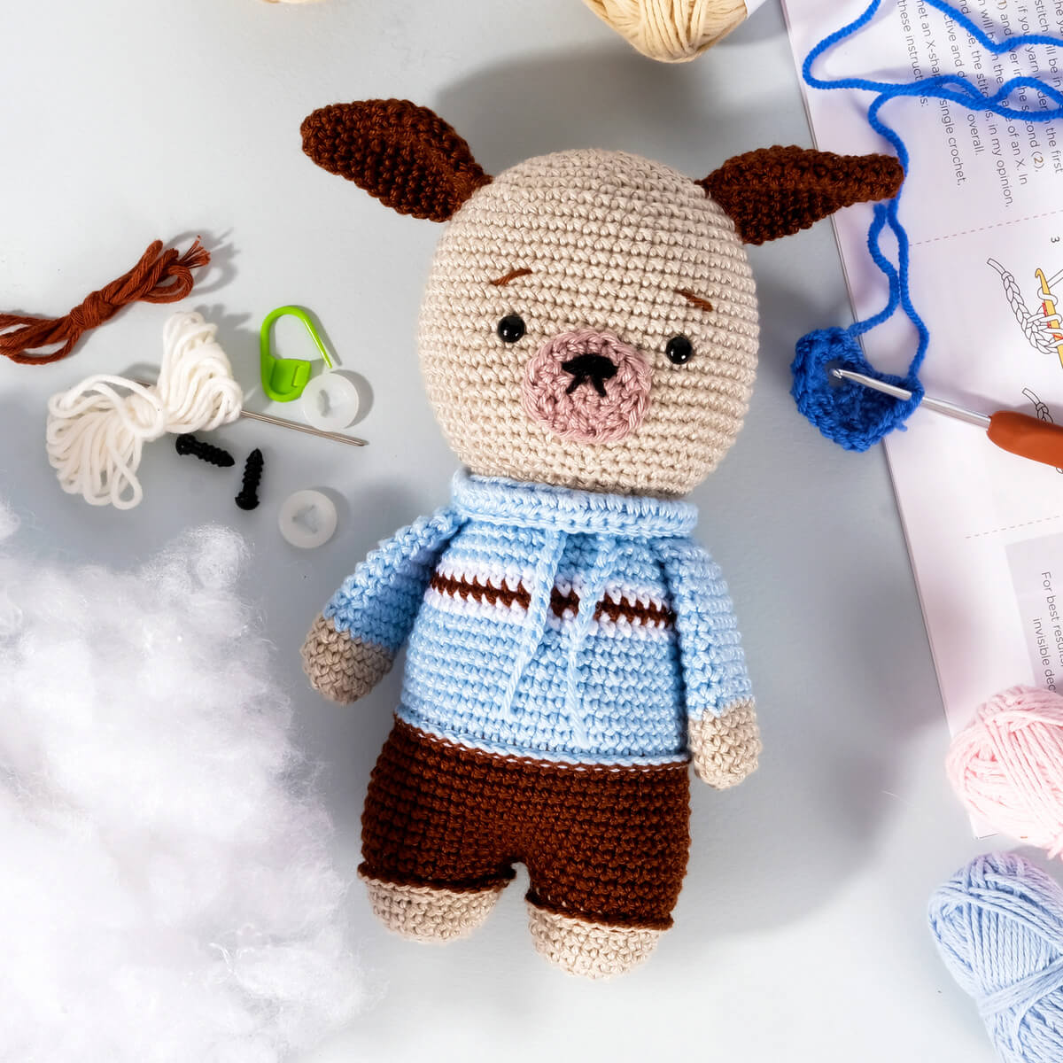 An Amigurumi Subscription Box featuring a crocheted dog doll in a blue and brown outfit lies on a crafting table surrounded by yarn, stuffing material, crochet hooks, and other crafting supplies.