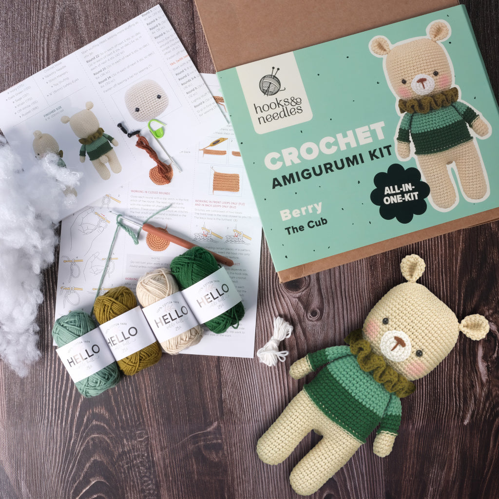 Amigurumi Subscription Box with yarn, stuffing, instructions, and a finished bear toy. The kit box is labeled "Hooks & Needles: CROCHET AMIGURUMI KIT – Berry The Cub".