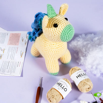 An Amigurumi Subscription Box featuring a crocheted yellow unicorn with a green snout and blue mane is surrounded by crafting materials including yarn, stuffing, a crochet hook, and a crochet pattern.