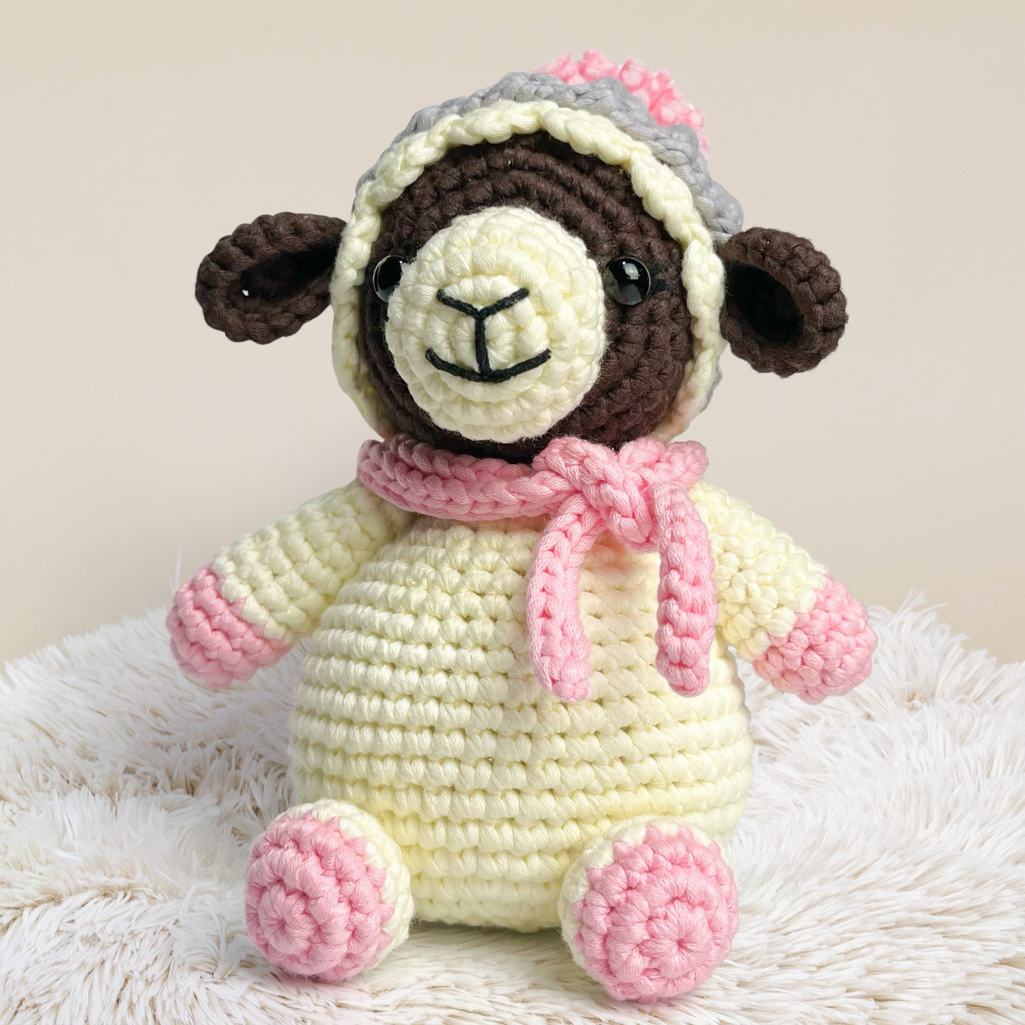 Wooly The Sheep Crochet Pattern features an adorable amigurumi sheep with a brown face, yellow body, pink hooves, and a pink scarf, displayed on a fluffy white surface against a neutral background.