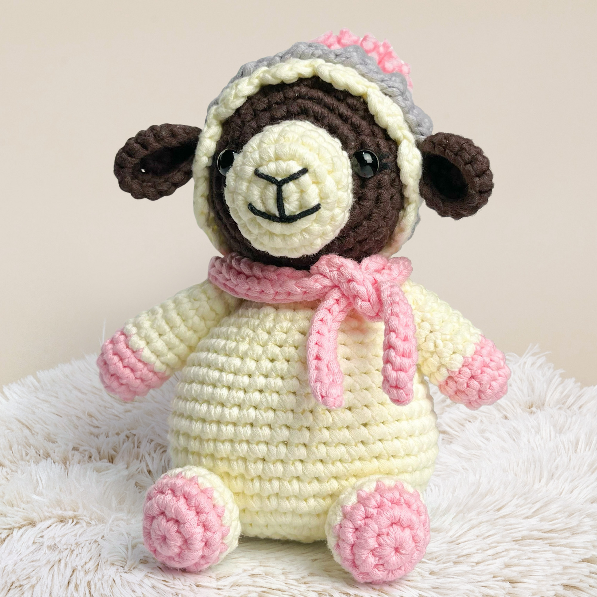 Wooly The Sheep Crochet Pattern features an adorable amigurumi sheep with a brown face, yellow body, pink hooves, and a pink scarf, displayed on a fluffy white surface against a neutral background.